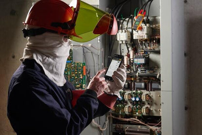 En elektriker i beskyttelsesudstyr bruger Fluke 377 FC tangamperemeter og smartphone foran en el-tavle.